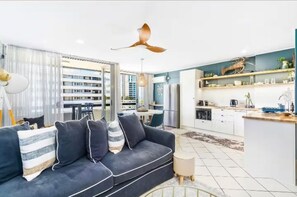 Una sala de estar moderna con un sofá azul oscuro, una zona de comedor y una cocina con armarios blancos y salpicadero azul.