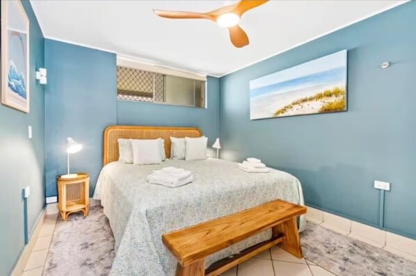 Un dormitorio con cama, ventilador de techo, banca, mesita de noche y un cuadro de una escena de playa.