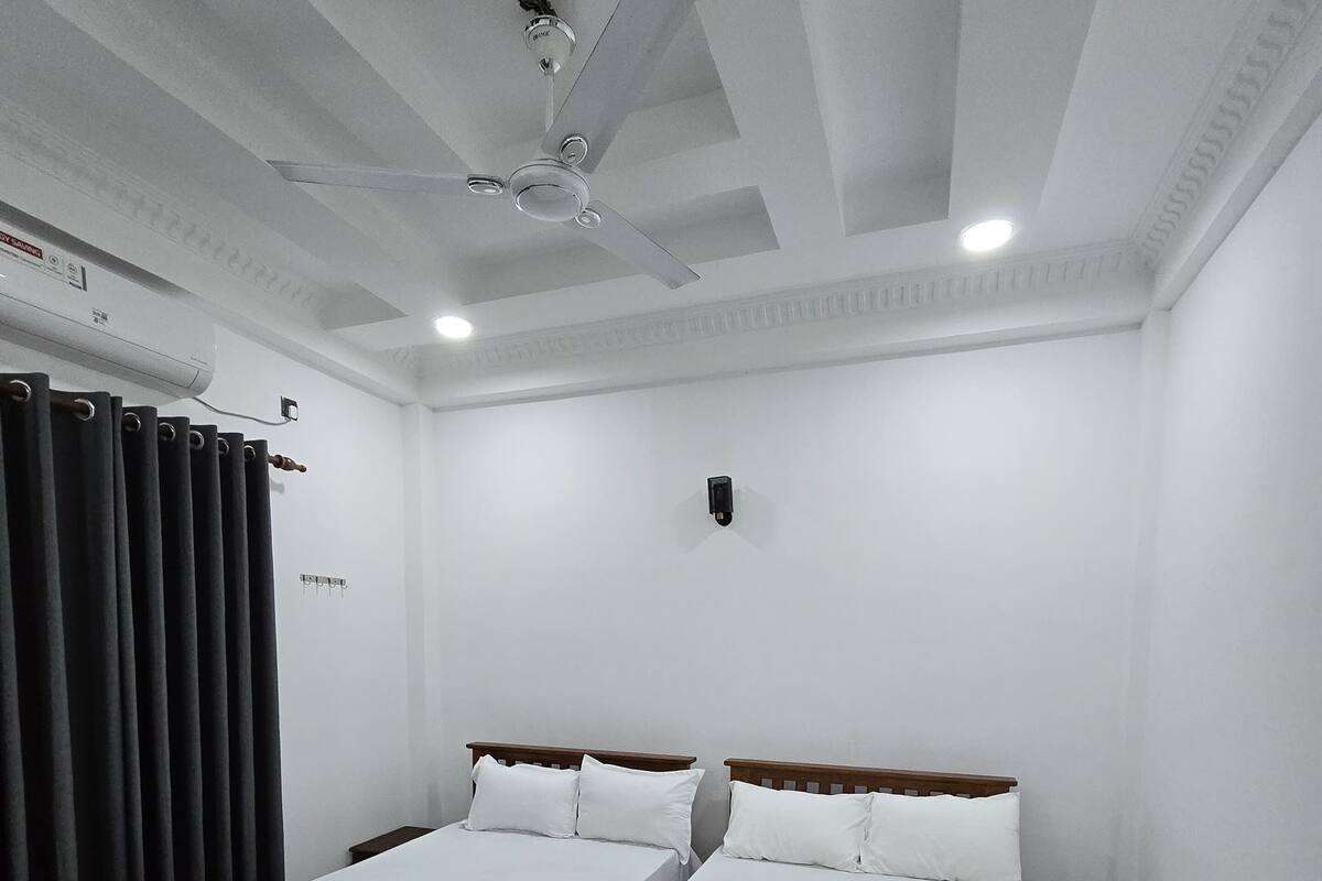 A hotel room with two beds, a ceiling fan, an air conditioning unit, a small bedside table, and a nightstand.