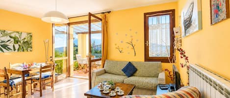 A bright living room with a sofa, a dining table, and chairs. There is a view through a glass door to an outdoor area.