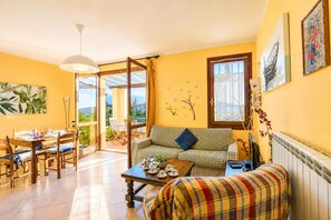Una sala luminosa con un sofá, una mesa de comedor y sillas. Se ve un espacio exterior a través de un vidrio.