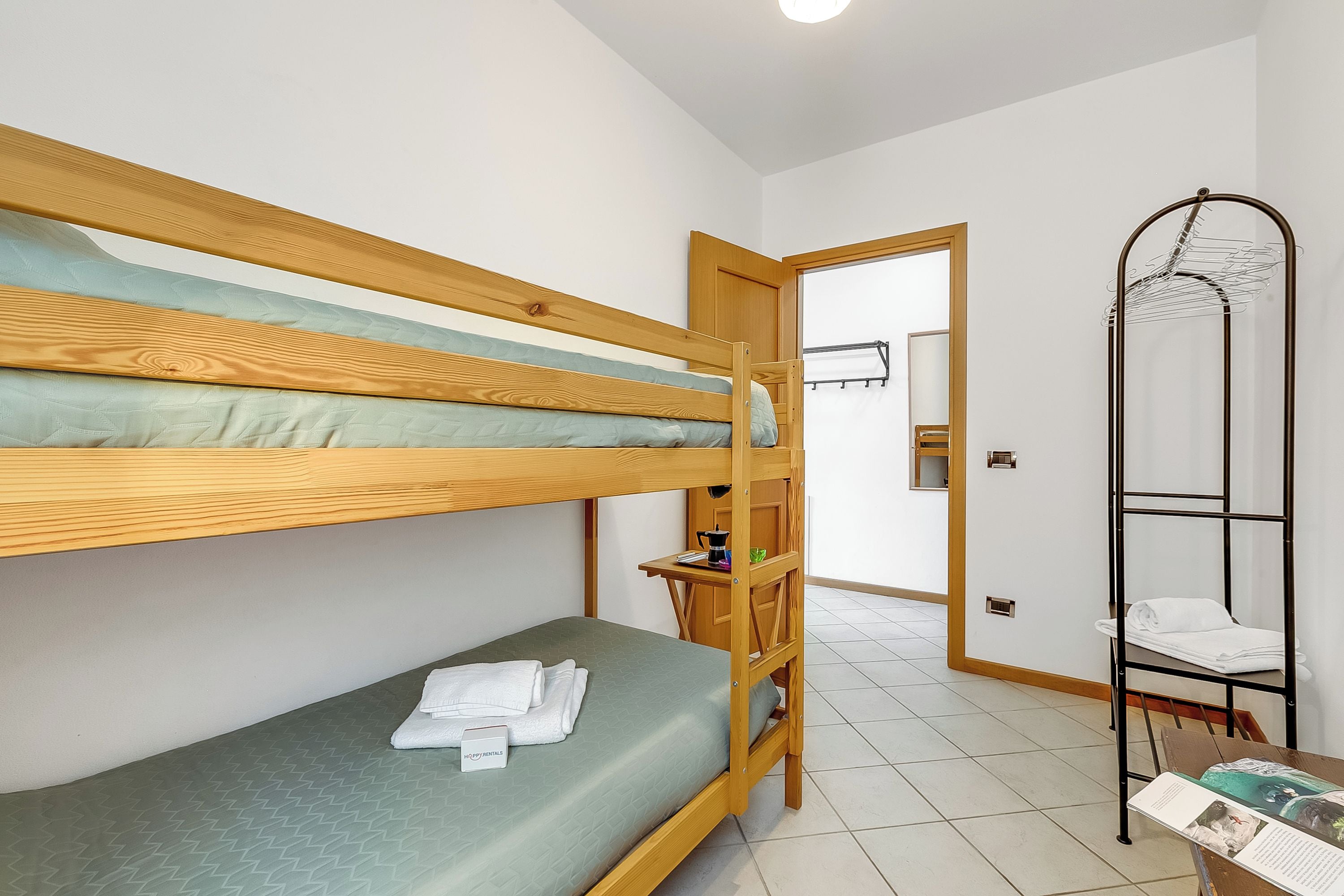 Ein Zimmer mit Etagenbetten, einem Holztisch, einem Wäscheständer aus Metall und einer Tür zu einem weiteren Raum.