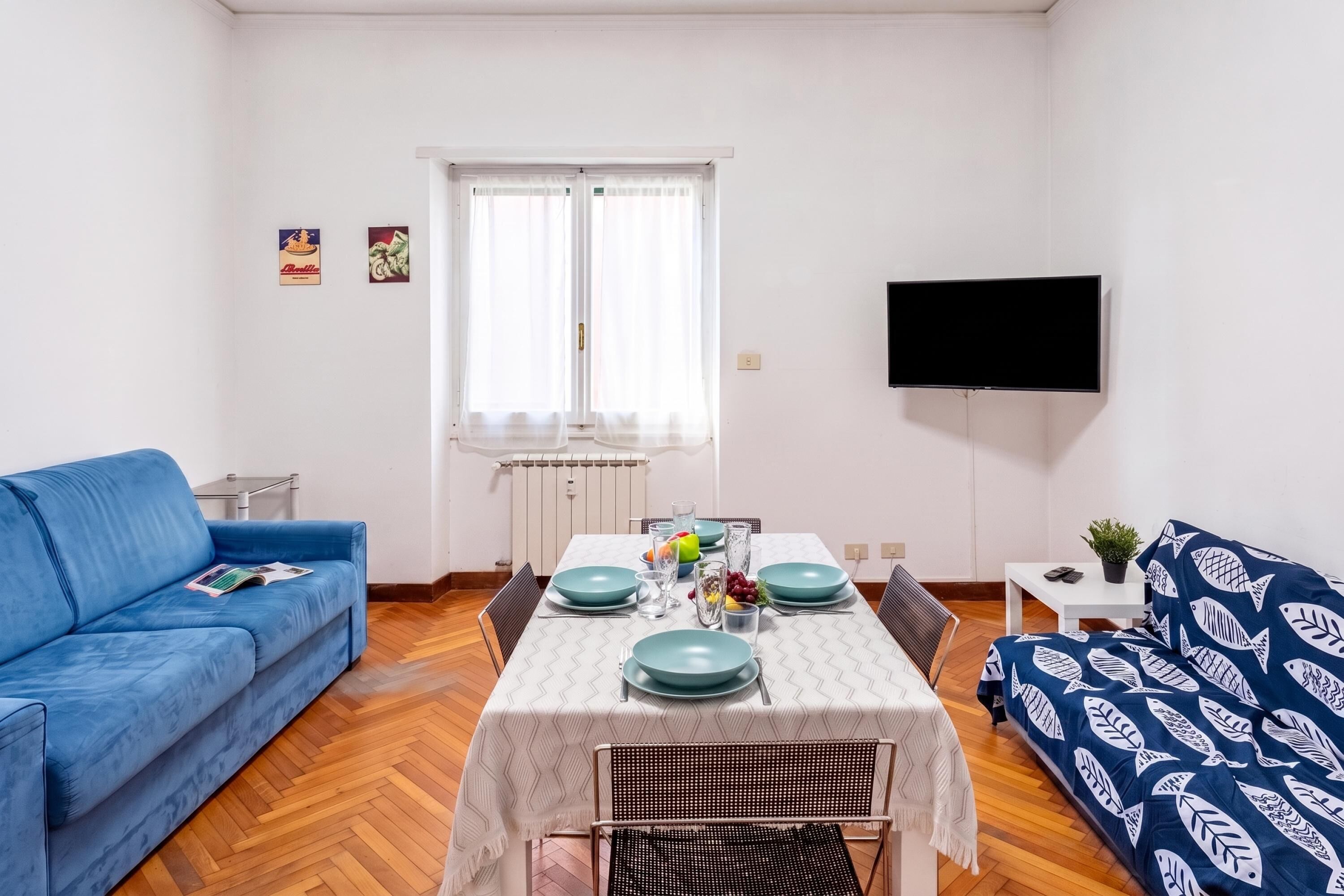 A modern living room with a blue sofa, a dining table set with blue plates, and a flat-screen TV mounted on the wall.