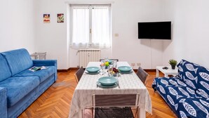 A modern living room with a blue sofa, a dining table set with blue plates, and a flat-screen TV mounted on the wall. - House Rondinella Genova City Center (Genoa)
