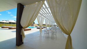 A covered outdoor seating area with white chairs and tables, a pool in the background, and white curtains tied at the corners. - Agricola Chiani (Melendugno)
