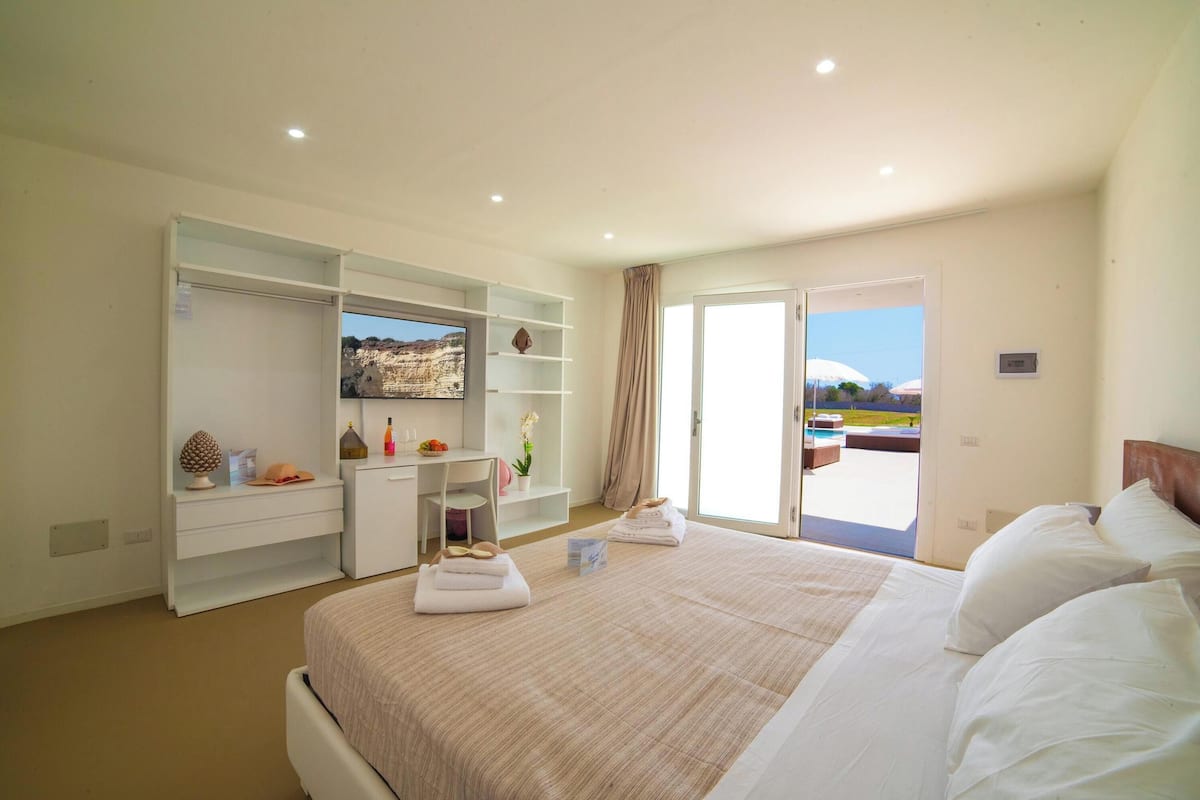 A hotel room with a bed, a desk, a wardrobe, and a view of a pool and landscape.