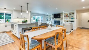 Een moderne keuken met witte kasten, een eethoek met een houten tafel en stoelen, en een zithoek met een open haard en planken.