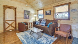 A cozy cabin interior with a wooden floor, a brown leather sofa, a wooden coffee table, and a blue rug. - Horse Farm Retreat w/ Pond View Near Louisville (La Grange)