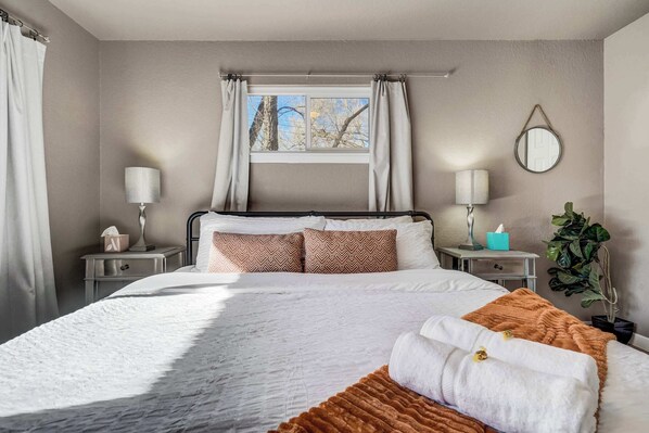 A bedroom with a bed, two bedside tables, a window with curtains, a mirror, and a potted plant.