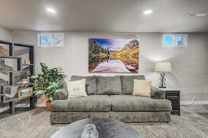 A living room with a grey sofa, a coffee table, a potted plant, a painting of a mountain landscape, and a lamp.