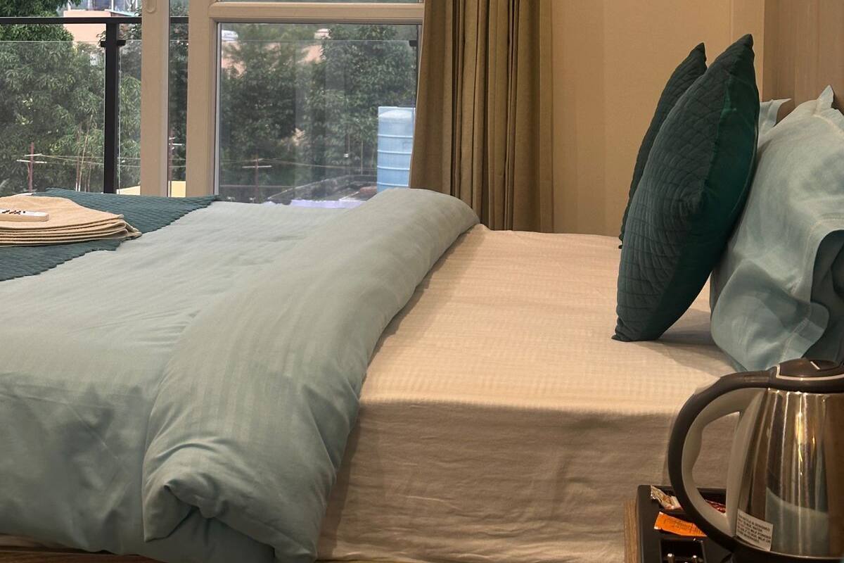A bedroom with a large window, a bed with pillows, a bedside table with a kettle, and a view of a hill with a building.