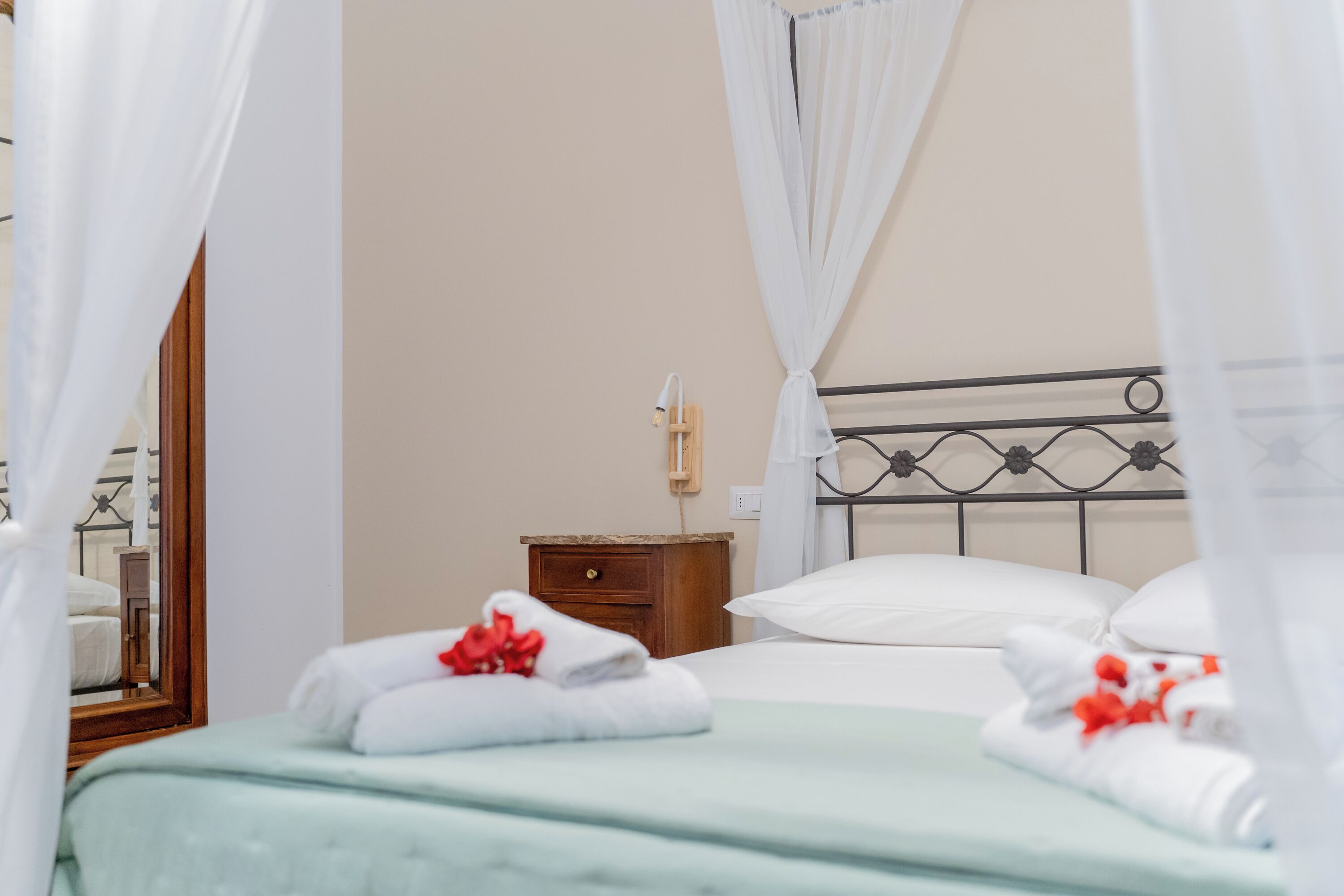 Cama bem arrumada com roupa de cama branca e detalhes vermelhos, mesa de cabeceira de madeira com luminária e cabeceira de metal.