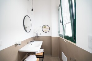 Une petite salle de bain avec un lavabo blanc, deux miroirs, un porte-serviettes et une fenêtre avec des stores.