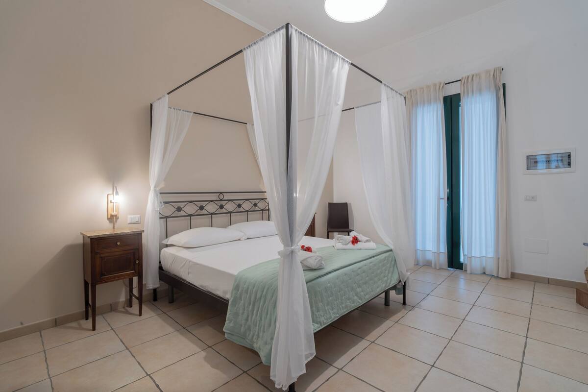 Un lit à baldaquin avec une couvre-lit verte, une table de chevet avec une lampe et une porte avec des rideaux.