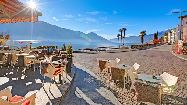 An outdoor café with tables and chairs, a clear view of the sea and mountains, and a sunny sky. - O Lac Ascona With Garden (Ascona)