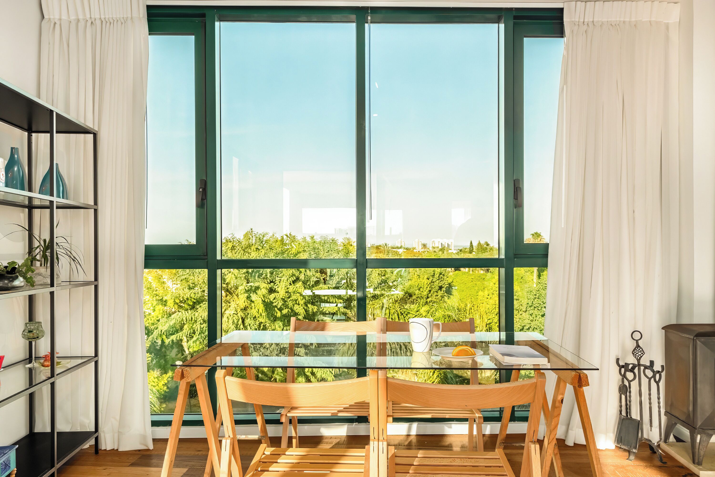 A dining area with a glass table and chairs, a bookshelf, and a view of greenery through large windows.
