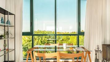 A dining area with a glass table and chairs, a bookshelf, and a view of greenery through large windows.
