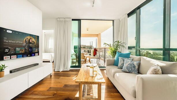 A modern living room with a flat-screen TV, wooden coffee table, white sofa, and large windows offering a view of the city. -  Caesarea Sea Elegant Stay by Sea N' Rent (Hof HaCarmel)