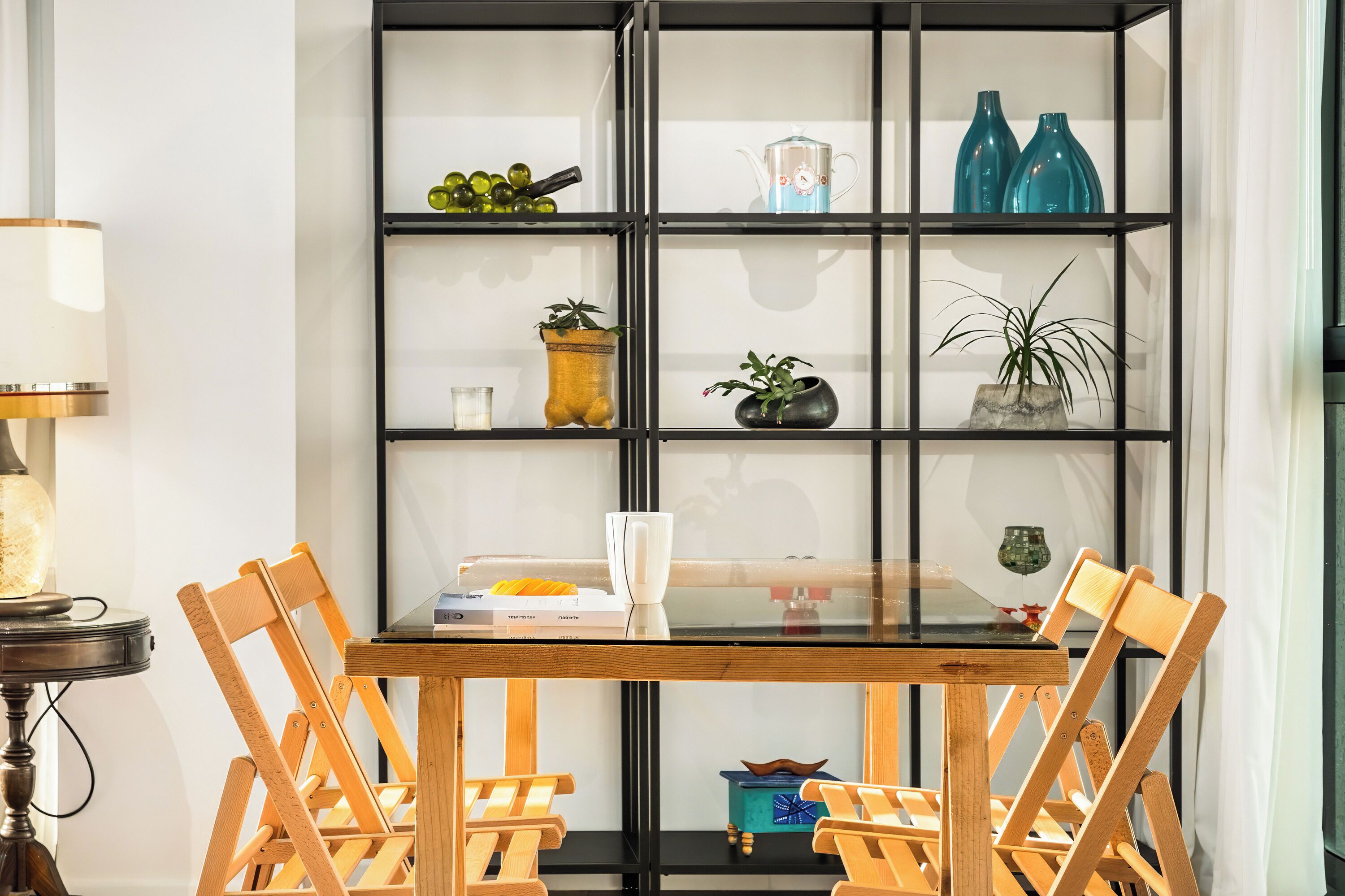 A dining area with a glass-top table, wooden chairs, and a black metal shelving unit with decorative items.