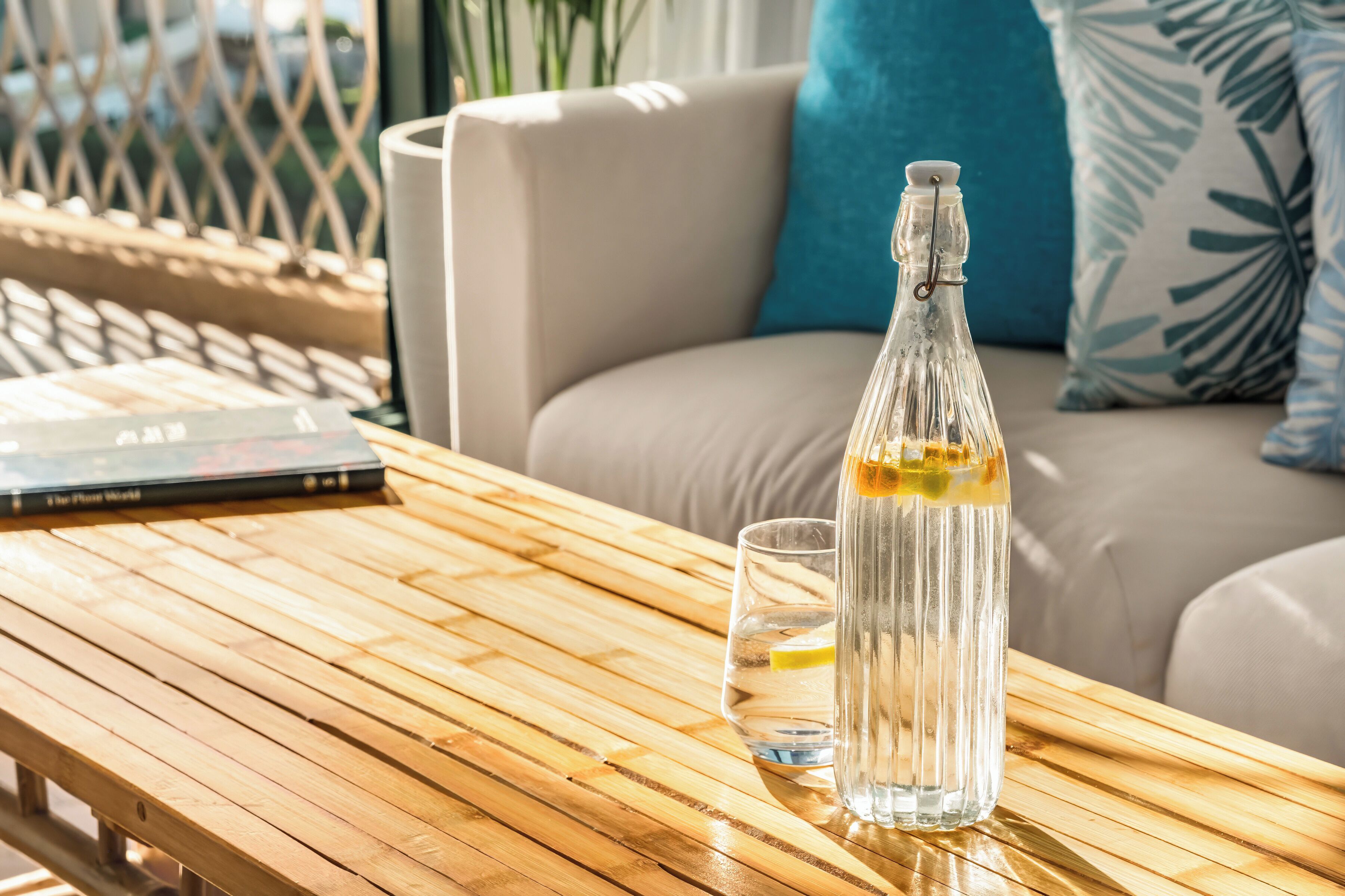 Una botella de vidrio transparente con una rodaja de limón sobre una mesa de madera, un vaso de agua y un libro titulado 