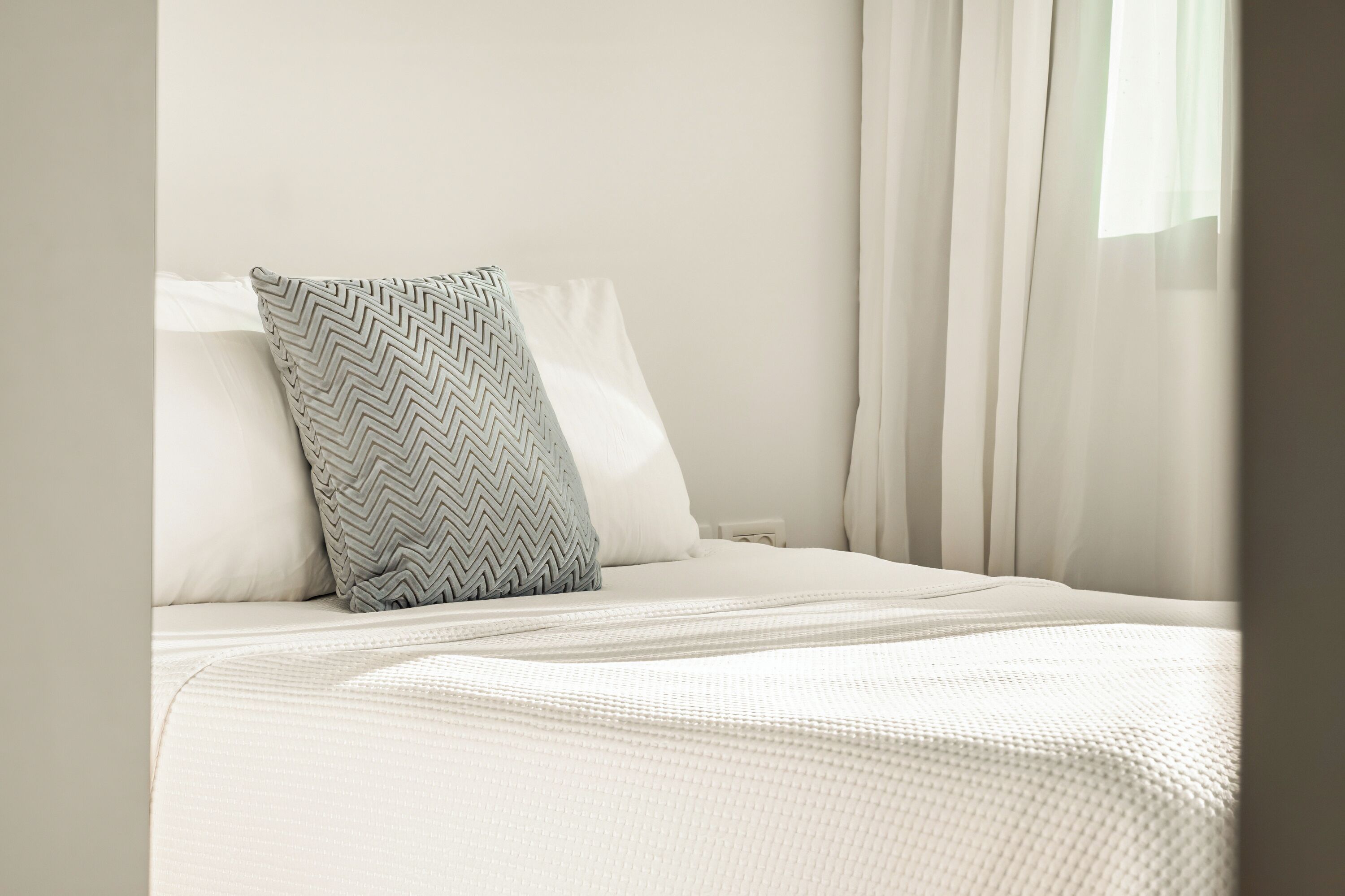 A neatly made bed with a patterned pillow, white bedding, and sheer curtains.