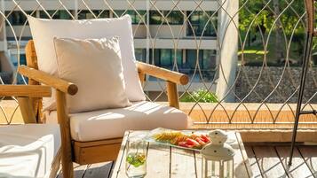 Una silla de madera con un cojín blanco, una mesa de vidrio con un plato de fruta y un farol, y una vista de un edificio a través de una cerca de malla metálica.