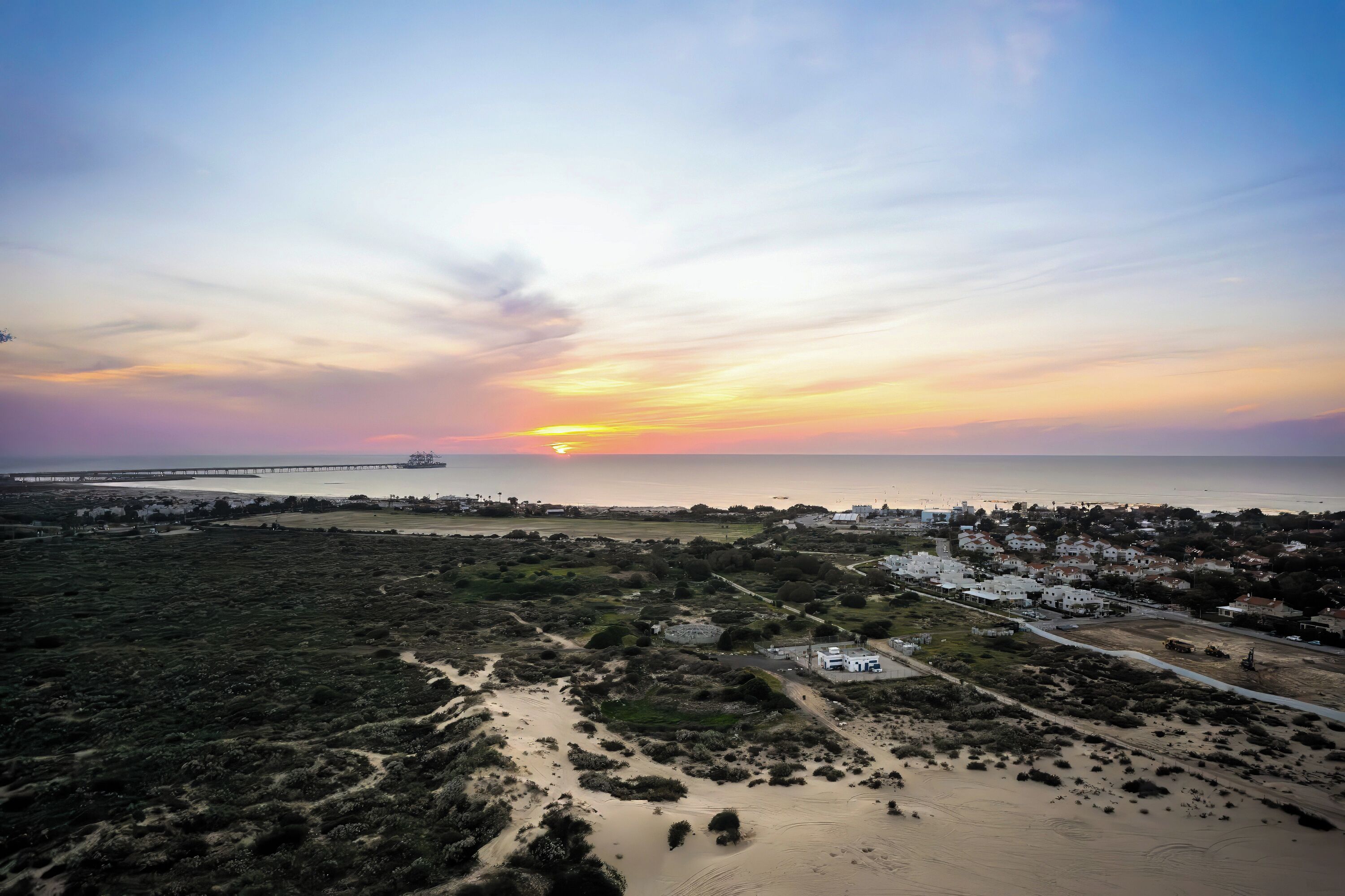 An aerial view of a coastal area with a sunset, a bridge, and a residential area with white buildings.