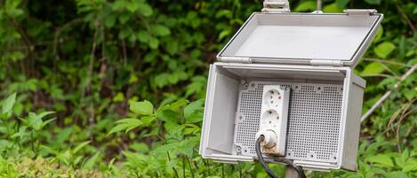 A metal electrical box with a hinged cover, mounted on a post with a cable connected to it, set against a backdrop of greenery.