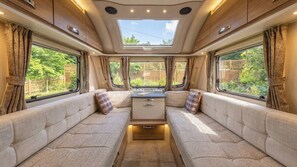 Interior acogedor de una caravana, con cocina central, asientos acolchados y ventanas amplias con vistas a la vegetación.