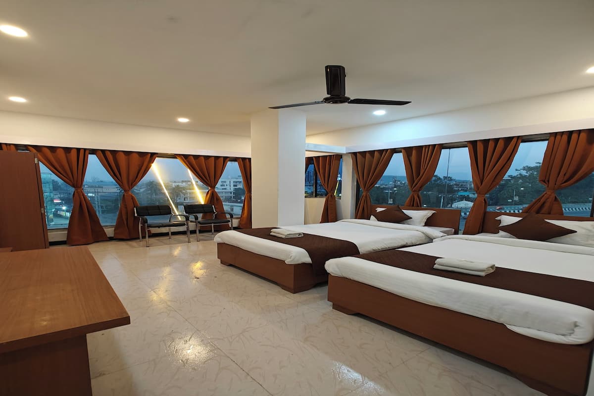 A modern hotel room with two large beds, a desk, and a chair. The room has large windows with curtains, a ceiling fan, and a city view.