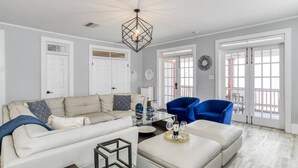 A spacious living room with a beige sofa, a blue armchair, and a glass coffee table. There is a geometric chandelier, white doors, and a view through large windows. - 20 Hypolita Street (St. Augustine)