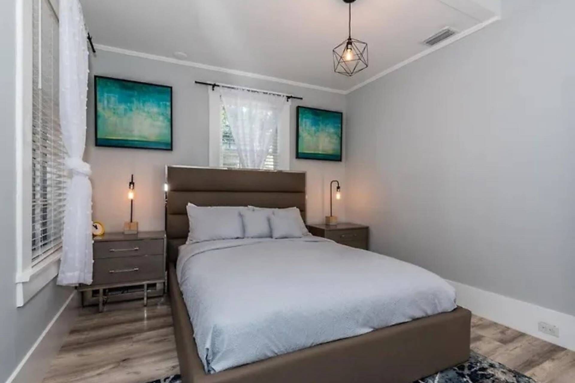 Een moderne slaapkamer met een groot bed, twee nachtkastjes, een geometrisch hanglamp en twee abstracte schilderijen aan de muur.