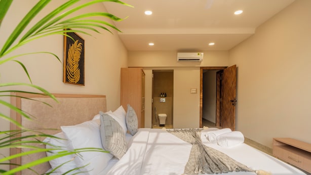 A hotel room with a bed, a bedside table, a nightstand, a wall-mounted air conditioner, and a door leading to a bathroom. - ELIVAAS Kamerios Regalia 2BHK Apartment (Marra)