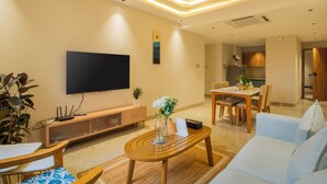 A modern living room with a flat-screen TV, wooden furniture, a dining area, and a kitchen in the background. - ELIVAAS Kamerios Regalia 2BHK Apartment (Marra)