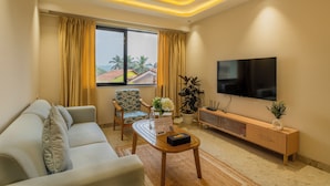A modern living room with a sofa, a coffee table, a TV, and a view of palm trees. - ELIVAAS Kamerios Regalia 2BHK Apartment (Marra)