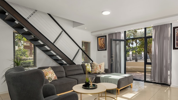 A modern living room with a staircase, a sofa set, a coffee table, and a view through large windows. - Modern 3-Bed Home , Canal Views & Parking (Cape Town)