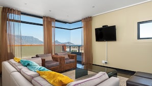 A modern living room with a sofa, armchairs, and a glass coffee table. A flat-screen TV is mounted on the wall, and the room has large windows with a view of a mountain and cityscape. - Modern 3-Bed Home , Canal Views & Parking (Cape Town)