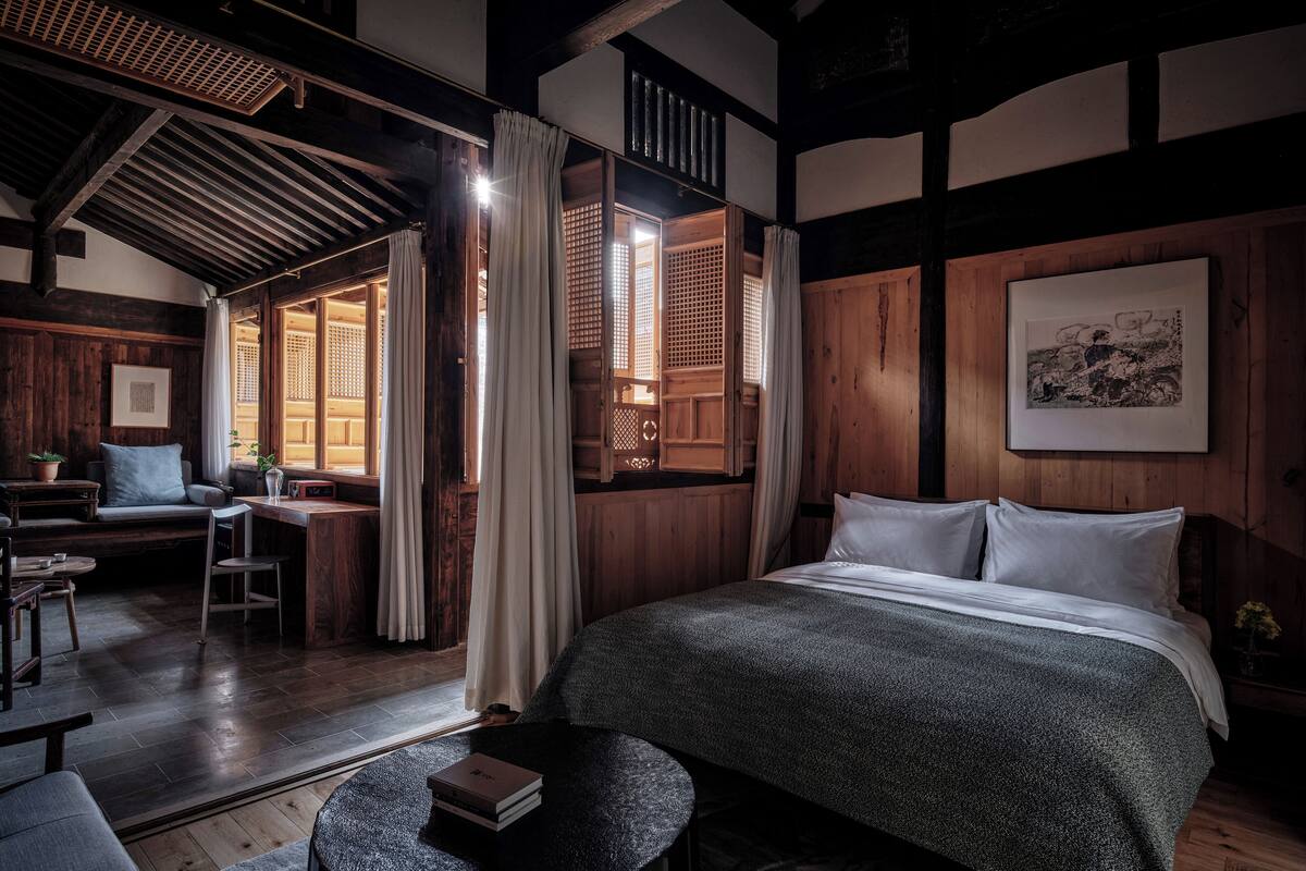 A traditional Japanese-style room with tatami flooring, a low wooden desk, a bed with a dark blanket, and a large window with wooden lattice.
