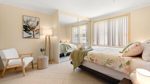 A bedroom with a bed, a chair, a bedside table, a lamp, and a mirror.