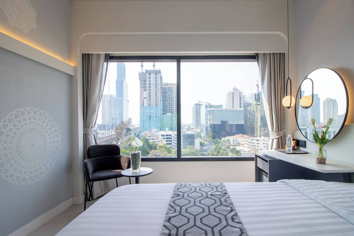 A hotel room with a large bed, a small table, a chair, a desk, a mirror, and a view of a cityscape through a large window.
