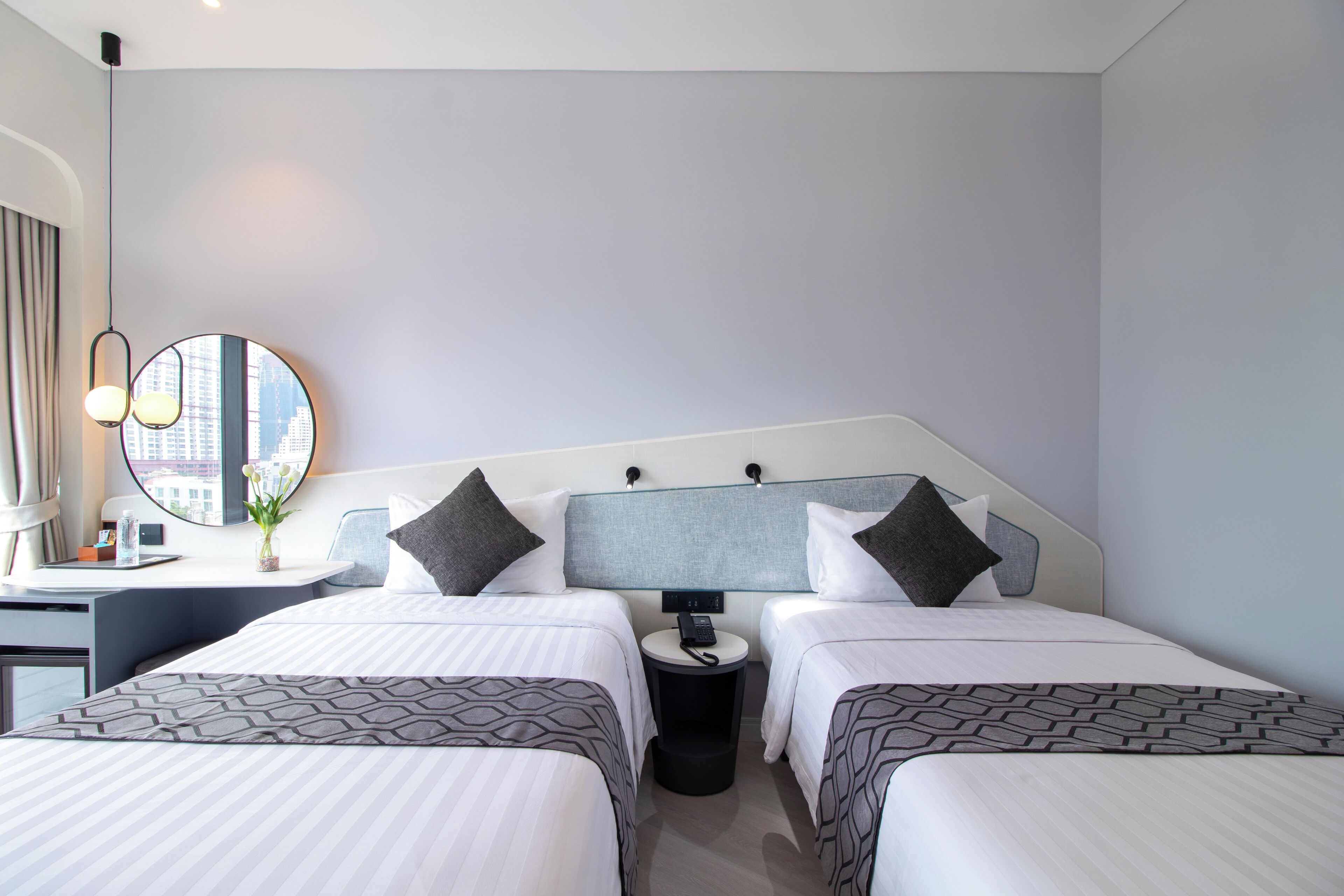 Habitación de hotel con dos camas, un escritorio, una silla, un espejo redondo y una pequeña mesa con un jarrón de flores.