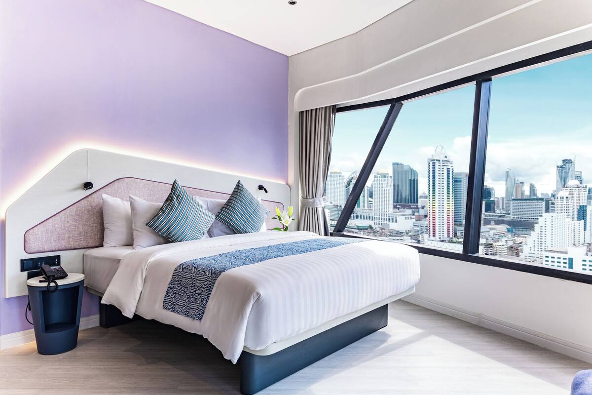 A modern bedroom with a city view, a large bed, bedside table, and a trash bin.