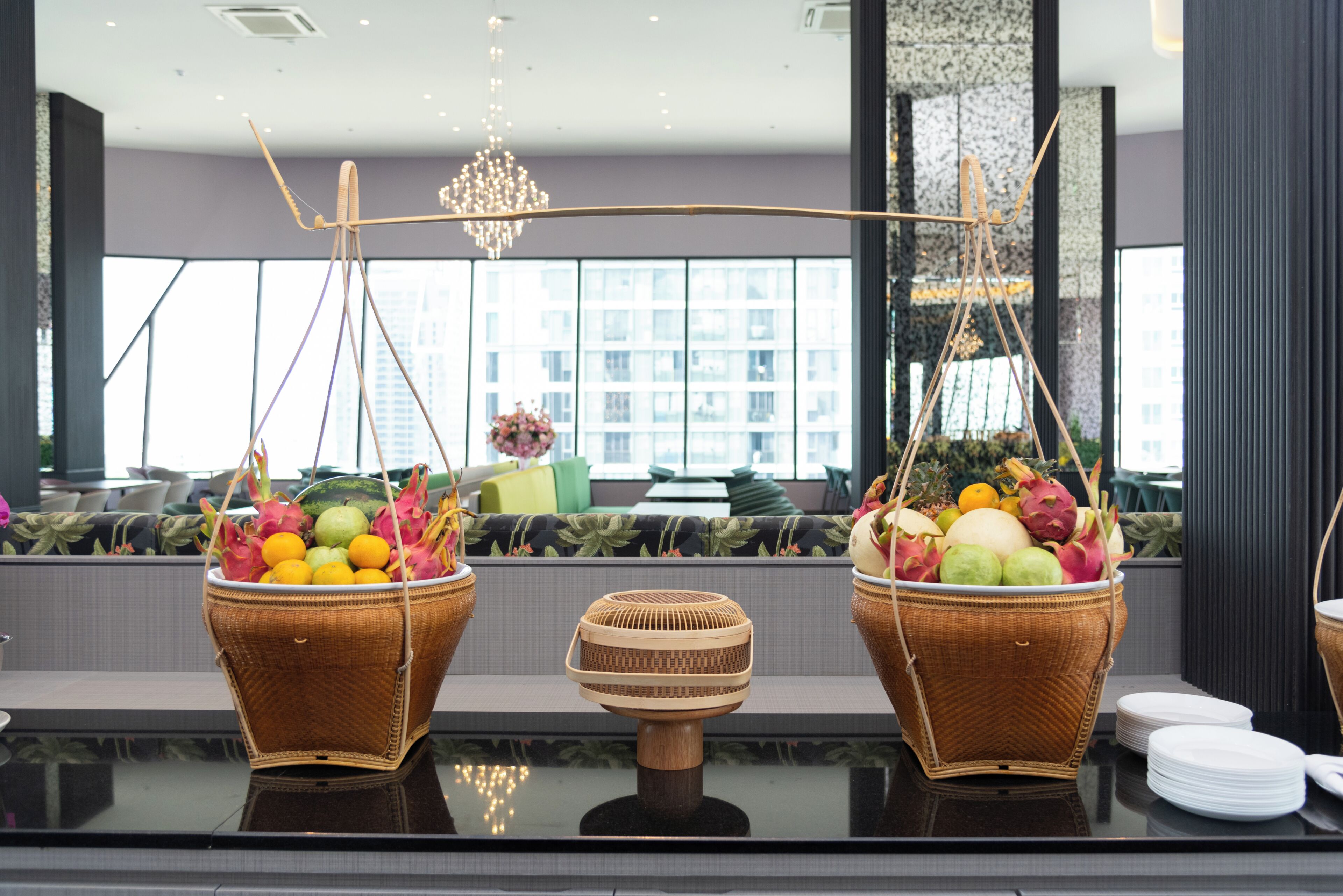 Two wicker fruit baskets on a reflective surface with a modern interior, including a chandelier and large windows.
