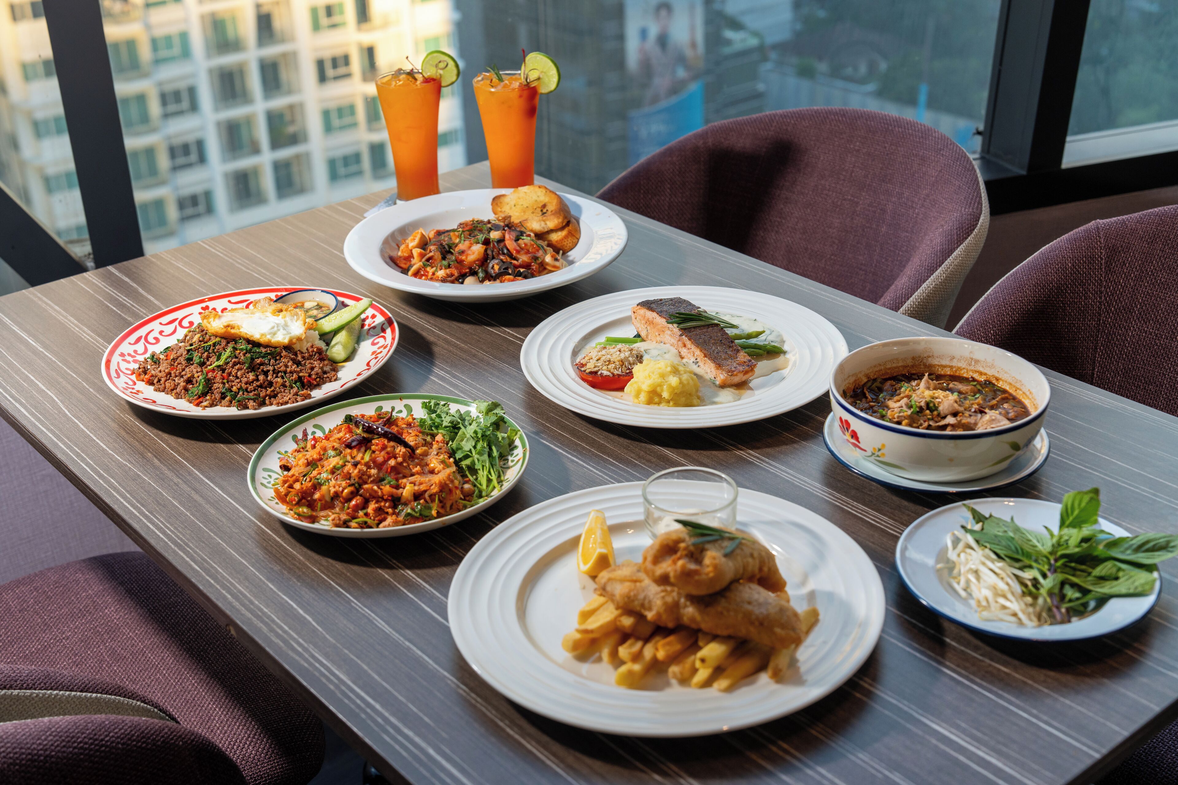 A table set with various dishes including seafood, pasta, and a salad, with a city view in the background.
