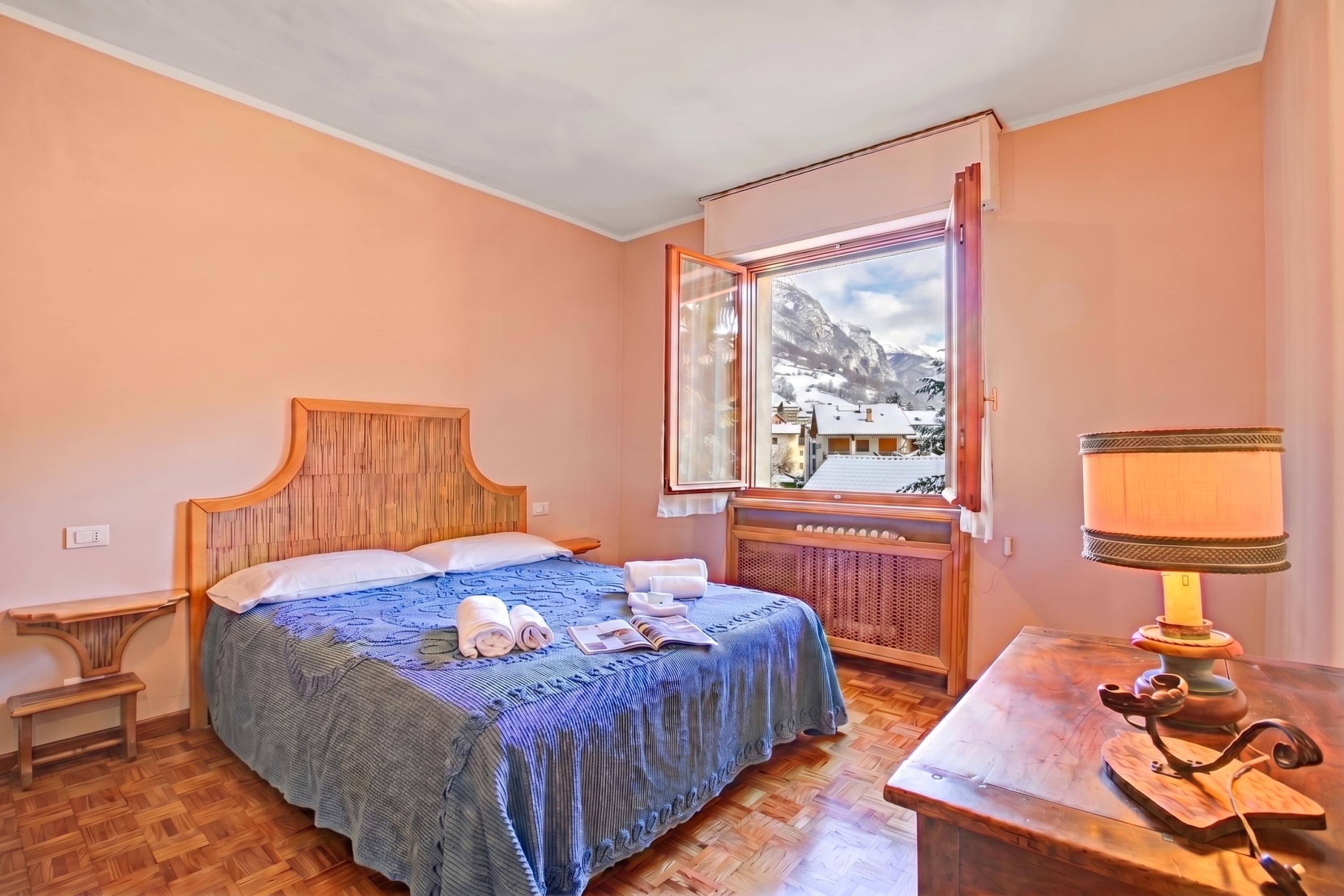 A bedroom with a wooden bed, a window with a view of a snowy mountain, and a bedside table with a lamp.
