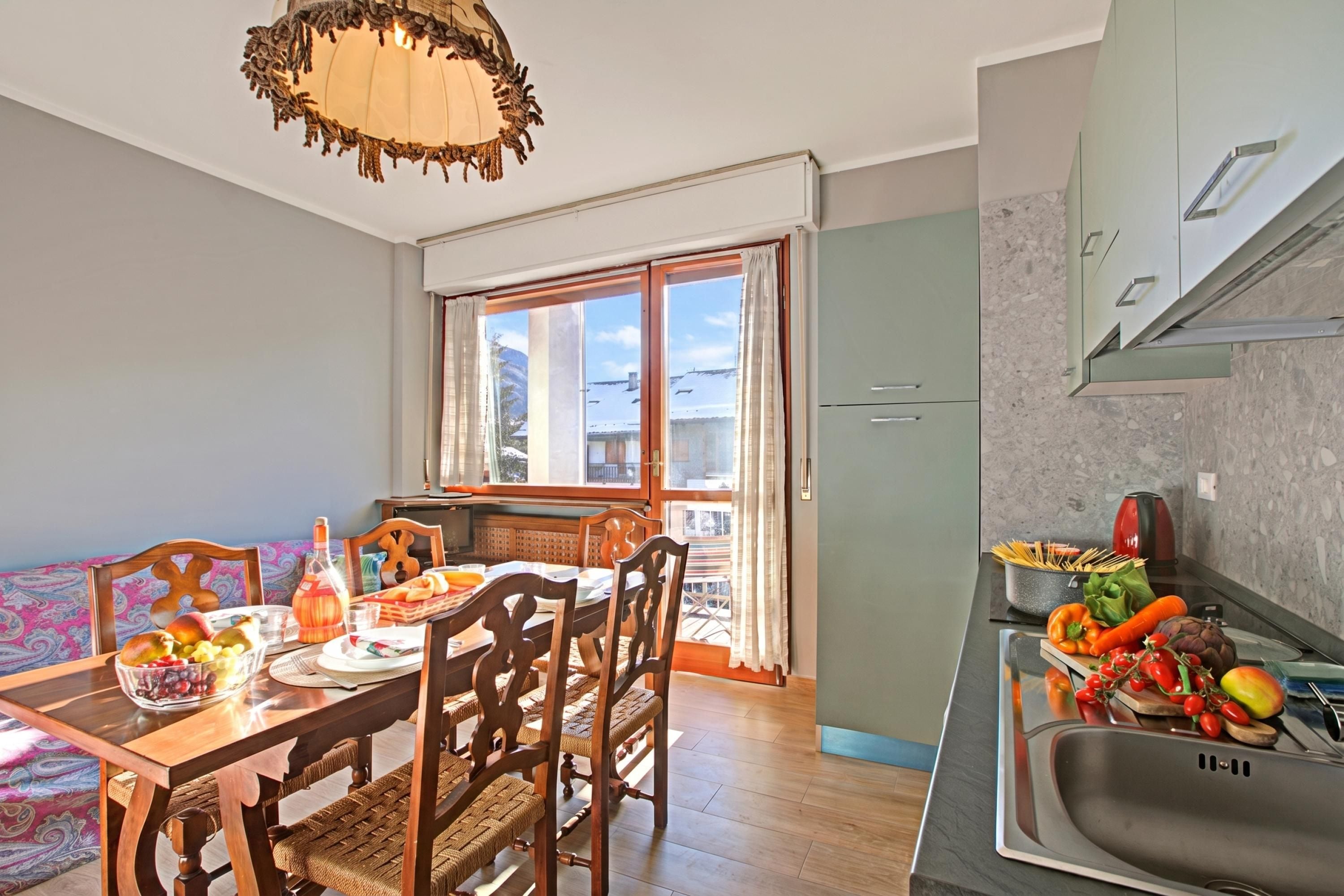 A dining area with a wooden table and chairs, a kitchen with a sink and stove, and a window with curtains.