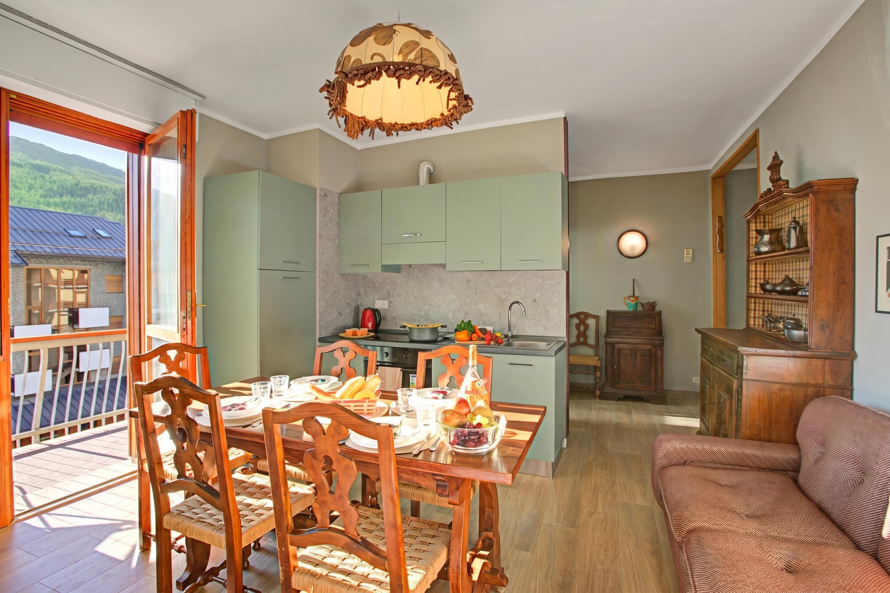 Ein Essbereich mit Holzkohlen und einem Tisch für zwei Personen, eine Küche mit grünen Schränken und ein Balkon mit Bergblick.