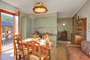 A dining area with wooden chairs and a table set for two, a kitchen with green cabinets, and a balcony with a view of the mountains. - La Casa Di Felice (Bardonecchia)