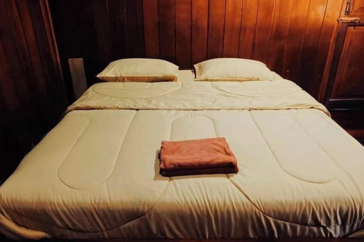 Un lit simple avec une couvre-lit beige et deux oreillers, une couverture marron pliée et des panneaux de bois aux murs.