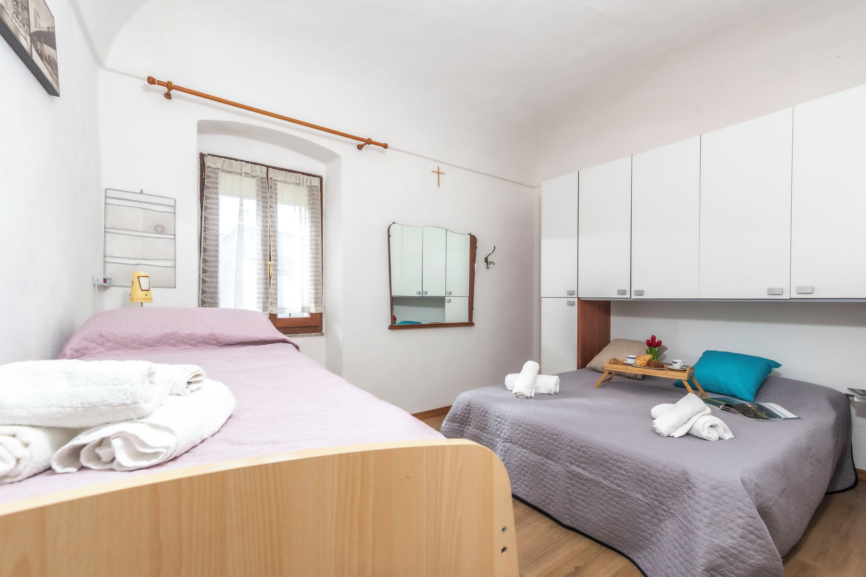A bedroom with two beds, a wooden headboard, a window with curtains, a mirror, a cross on the wall, and a small table with towels and a tray.
